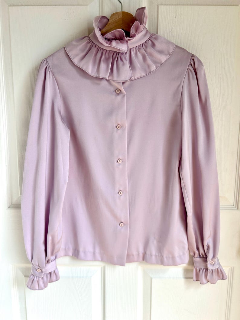 May include: A light purple blouse with a ruffled collar and cuffs. The long-sleeved shirt has a button-down front and is hanging on a wooden hanger. The fabric appears to be a soft, flowing material.