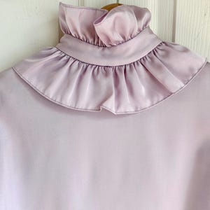 May include: A light purple blouse with a high, ruffled collar. The blouse is made of a smooth, silky material and hangs on a wooden hanger. The ruffled collar adds a vintage touch to the garment.