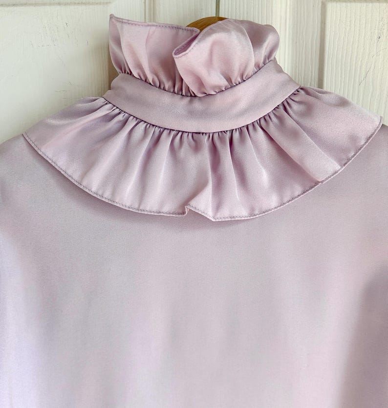 May include: A light purple blouse with a high, ruffled collar. The blouse is made of a smooth, silky material and hangs on a wooden hanger. The ruffled collar adds a vintage touch to the garment.