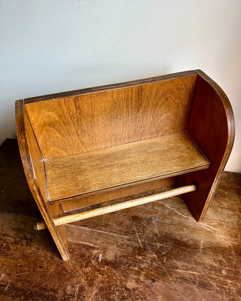 May include: A wooden paper towel holder with a shelf. The holder is made of brown wood and has a natural finish. The shelf is located above the paper towel roll.