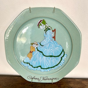 Op de afbeelding: Een achthoekig lichtgroen keramieken bord met een centrale illustratie van een persoon in een blauw-wit gestreepte jurk met groene randen, zittend op een stoel met een klein kind in een gele outfit. De tekst Anna Karenina staat onderaan gedrukt.