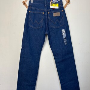 Puede incluir: Vaqueros Wrangler Cowboy Cut azul oscuro colgados de una percha de madera. Los vaqueros tienen un dise&ntilde;o cl&aacute;sico de cinco bolsillos, con el logo de Wrangler en el bolsillo trasero y una etiqueta con el nombre de la marca. Los vaqueros son una talla 86cm x 81cm.
