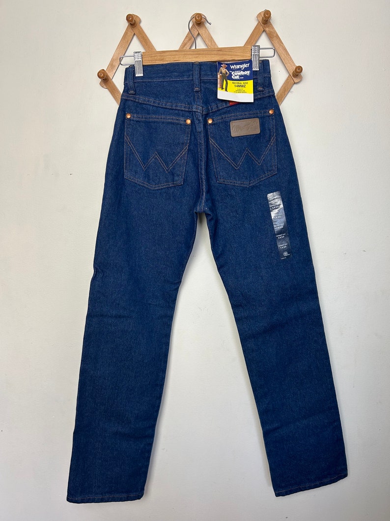 Puede incluir: Vaqueros Wrangler Cowboy Cut azul oscuro colgados de una percha de madera. Los vaqueros tienen un dise&ntilde;o cl&aacute;sico de cinco bolsillos, con el logo de Wrangler en el bolsillo trasero y una etiqueta con el nombre de la marca. Los vaqueros son una talla 86cm x 81cm.