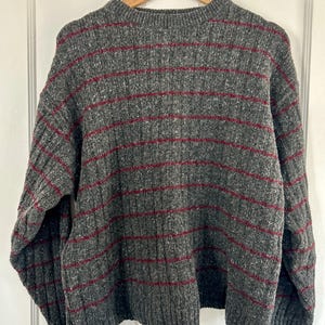May include: A gray sweater with horizontal burgundy stripes. The sweater has a ribbed texture and a crew neck. It is hanging on a wooden hanger against a white background. The sweater appears to be made of a thick, warm material.