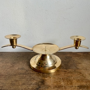 Puede incluir: Un candelero de latón dorado con dos brazos y una base central. El candelero tiene un diseño simple y elegante.
