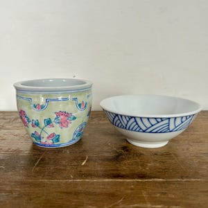 May include: Two ceramic bowls on a wooden surface. One is a small, round bowl with a white interior and a blue geometric pattern on the exterior. The other is a small, cylindrical bowl with a floral design.