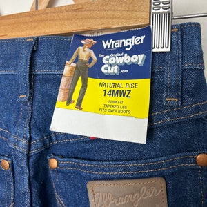Puede incluir: Primer plano de vaqueros Wrangler Cowboy Cut de mezclilla azul con tiro natural y corte ajustado. Los vaqueros tienen un parche de cuero con el logo de Wrangler y una etiqueta amarilla con informaci&oacute;n del producto. Los vaqueros est&aacute;n colgados de una percha de madera.