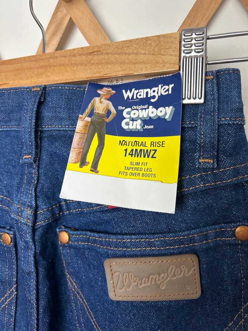 Puede incluir: Primer plano de vaqueros Wrangler Cowboy Cut de mezclilla azul con tiro natural y corte ajustado. Los vaqueros tienen un parche de cuero con el logo de Wrangler y una etiqueta amarilla con informaci&oacute;n del producto. Los vaqueros est&aacute;n colgados de una percha de madera.