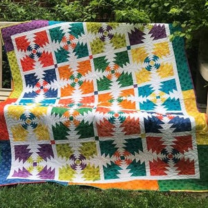 Könnte beinhalten: Eine farbenfrohe Quiltdecke mit einem geometrischen Muster aus Ananas. Die Decke besteht aus Quadraten in verschiedenen Farben, darunter Rot, Orange, Gelb, Grün, Blau und Lila. Die Decke ist gefaltet und liegt auf einer Holzbank.