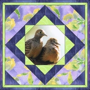 May include: Two brown and white doves with light brown feathers are perched together in a circular frame. The frame is surrounded by a quilt pattern of green, blue, and purple squares and triangles. The background is a blurry image of purple and green flowers.