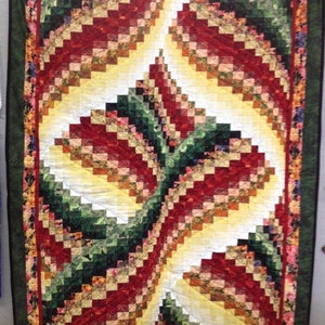 Op de afbeelding: Een handgemaakte quilt met een levendig, abstract ontwerp. De quilt heeft een centraal patroon van wervelende, rechthoekige blokken in tinten rood, geel en groen, omlijst door een bloemenrand en een donkergroene rand. Het is een decoratief textiel.
