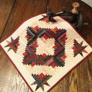 Puede incluir: Una pequeña colcha de color crema con un borde rojo. La colcha presenta un patrón de cabaña de troncos en tonos rojos, marrones y azules. La colcha se está cosiendo en una máquina de coser vintage.