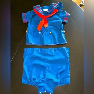 May include: A vintage blue sailor outfit for a baby or toddler. The top has a red collar and trim, with a red necktie. The matching shorts are a solid blue. The outfit has white buttons and is made of a soft fabric.