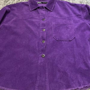 May include: A vibrant purple corduroy button-down shirt featuring a classic collar, a single chest pocket on the left side, and a raw, frayed hemline. The garment is secured with four visible buttons down the front placket and includes a Zara brand label.