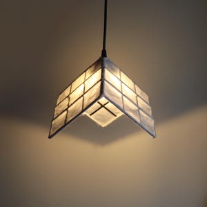 May include: A geometric pendant light fixture with a square, grid-like shade. The shade is a light, neutral color, possibly off-white or gray, and appears to be made of a textured material. The light is suspended from a black cord.