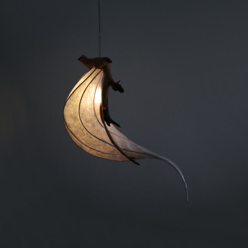 May include: A white paper lampshade with a natural wood branch design hangs from the ceiling. The lampshade is lit and casts a warm glow.