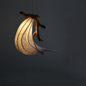 May include: A white paper lampshade with a natural wood branch attached. The lampshade is curved and has a flowing design. The light is on and illuminates the lampshade.