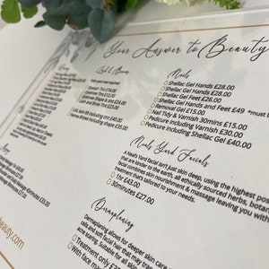 May include: A white acrylic sign with gold lettering lists beauty services and prices. The sign includes services like eyelash tinting, eyebrow shaping, shellac gel manicures, and dermaplaning. The sign also includes the text "Your Answer to Beauty".