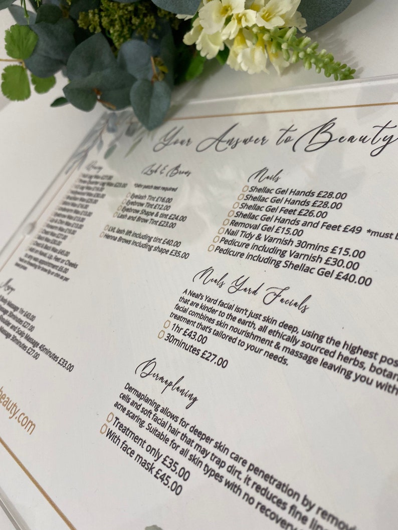 May include: A white acrylic sign with gold lettering lists beauty services and prices. The sign includes services like eyelash tinting, eyebrow shaping, shellac gel manicures, and dermaplaning. The sign also includes the text "Your Answer to Beauty".