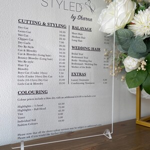 May include: A white acrylic sign with black text listing prices for hair cutting, styling, and coloring services. The sign is titled "Styled by Sharna".