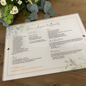 May include: A white sign with gold accents and a watercolor floral design lists beauty services and prices. The sign includes waxing, lash and brow services, nails, hand and foot treatments, and massages. The text "Your Answer to Beauty" is at the top of the sign.