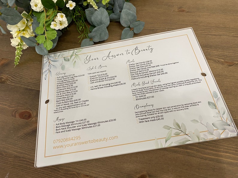 May include: A white sign with gold accents and a watercolor floral design lists beauty services and prices. The sign includes waxing, lash and brow services, nails, hand and foot treatments, and massages. The text "Your Answer to Beauty" is at the top of the sign.