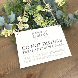 May include: White sign with black text that reads "Hannah Rebecca Aesthetics Do Not Disturb Treatment in Progress Please Contact 07506 623802 To Inform of Your Arrival". The sign is hanging from a string and is in front of a bouquet of white flowers.