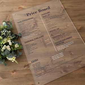 May include: A clear acrylic price board with black text listing prices for nail extensions, natural nail care, nail art, and extra services. The board includes prices for full sets, infills, shellac, gel powder, and SNS.