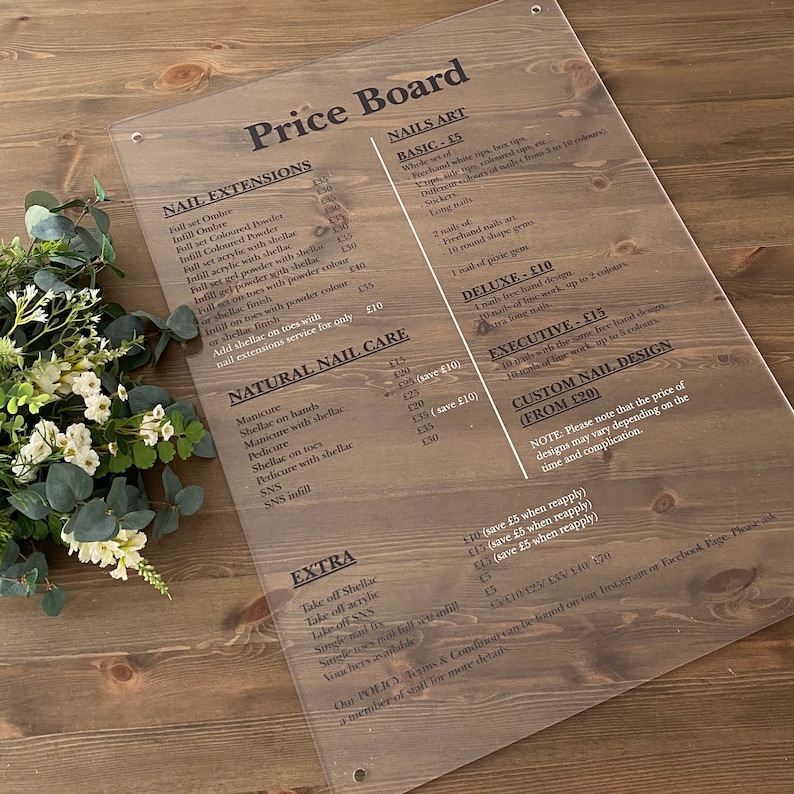 May include: A clear acrylic price board with black text listing prices for nail extensions, natural nail care, nail art, and extra services. The board includes prices for full sets, infills, shellac, gel powder, and SNS.