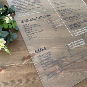 May include: A price board for a nail salon with a list of services and prices. The board is made of clear acrylic and has a wood grain background. The services include nail extensions, natural nail care, and extras. The prices are listed in pounds sterling.