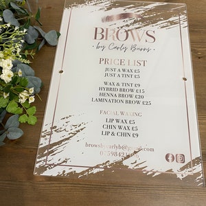 May include: A white acrylic sign with a pink and white abstract design. The sign advertises eyebrow services by Carly Burns. The price list includes services like waxing, tinting, hybrid brows, henna brows, and lamination. The sign also includes contact information and social media icons.