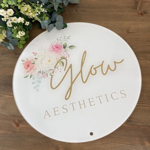 May include: A circular white sign featuring the words "Glow Aesthetics" in gold and grey. The sign is adorned with a floral design of pink and white roses and green foliage. A small hole is at the bottom.