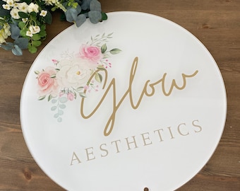 May include: A round white sign with the words "Glow Aesthetics" in gold and light gray. The sign is decorated with a floral arrangement of pink and white roses and green leaves. A small hole is at the bottom.