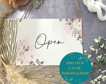May include: A white sign with the word "Open" written in black cursive script. The sign has a floral design with pink and green watercolor flowers. The sign is hanging from a string and has a teal circle with the text "This item can be personalised!" on the bottom right corner.