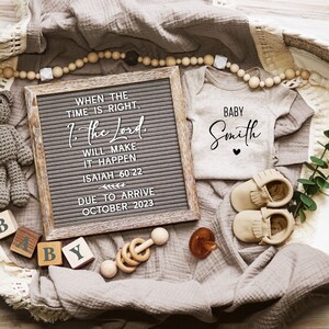 May include: A brown wicker basket filled with baby items, including a letter board with the quote "When the time is right, the Lord will make it happen. Isaiah 60:22" and the date "Due to arrive October 2023", a white onesie with the text "Baby Smith", a crocheted teddy bear, wooden blocks spelling "Baby", a wooden rattle, and a pair of brown moccasins.