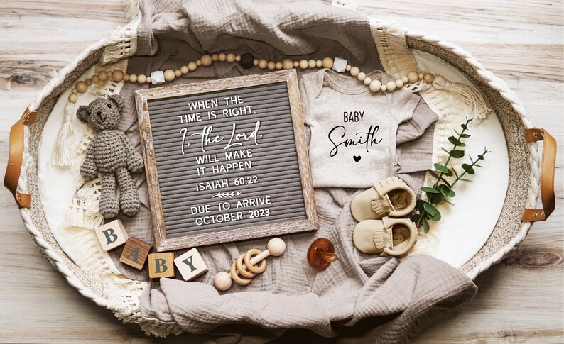 May include: A brown wicker basket filled with baby items, including a letter board with the quote "When the time is right, the Lord will make it happen. Isaiah 60:22" and the date "Due to arrive October 2023", a white onesie with the text "Baby Smith", a crocheted teddy bear, wooden blocks spelling "Baby", a wooden rattle, and a pair of brown moccasins.