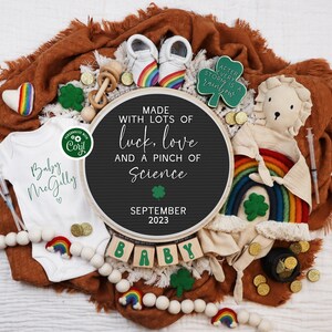May include: A baby announcement featuring a black letter board with the message "Made with lots of luck, love and a pinch of science" and "September 2023". The letter board is surrounded by a rainbow, shamrocks, gold coins, a baby onesie, and a stuffed lion.