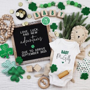 May include: A white onesie with the text "BABY McGilly" in green and black lettering. The onesie is surrounded by green shamrocks, wooden blocks that spell "BABY", and a wooden sign that reads "LOVE, LUCK, AND NEW adventures DUE TO ARRIVE SEPTEMBER 2023".