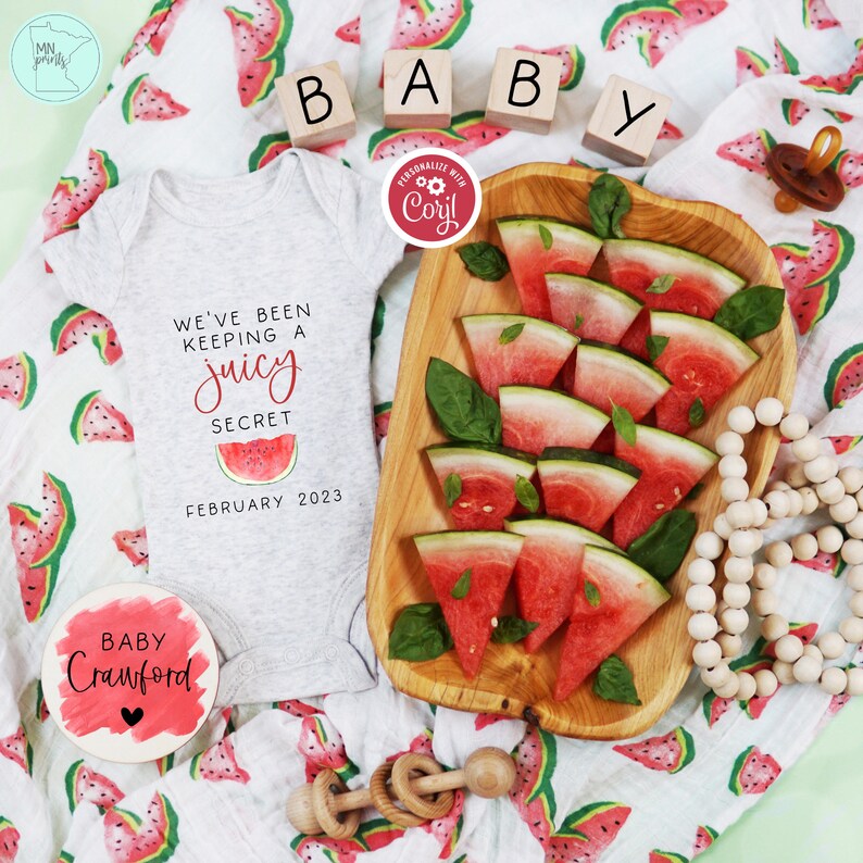 May include: A white onesie with the text "We've been keeping a juicy secret February 2023". A red and green watermelon print fabric is in the background. A wooden bowl with watermelon slices and a wooden bead necklace are also in the image.