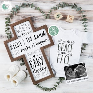 May include: A wooden cross with a white background and black text. The text reads "When the time is right, I the Lord will make it happen. Isaiah 60:22". Below the text is a heart and the words "Baby Huxley Nov. 2022". A white onesie with the text "All of God's Grace in one tiny face" and a black and white ultrasound image.