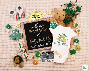 May include: A black letter board with the text "We found our pot of gold! Baby McGilly September 2023". A white onesie with a rainbow and the text "Little Lucky Charm". A small teddy bear, a shamrock, and gold coins are also in the image.