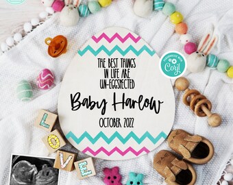 May include: A pink and blue chevron patterned wooden egg-shaped sign with the text "The best things in life are un-eggspected" and "Baby Harlow October 2022". The sign is surrounded by a white blanket, a pacifier, a rattle, a pair of baby shoes, and a few felt Easter bunny toys.