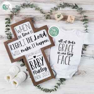 May include: A wooden cross with a white sign that reads "When the time is right, I the Lord will make it happen. Isaiah 60:22". Below the sign is a heart and the text "Baby Huxley Nov. 2022". A white onesie with the text "All of God's Grace in one tiny face" is next to the cross.