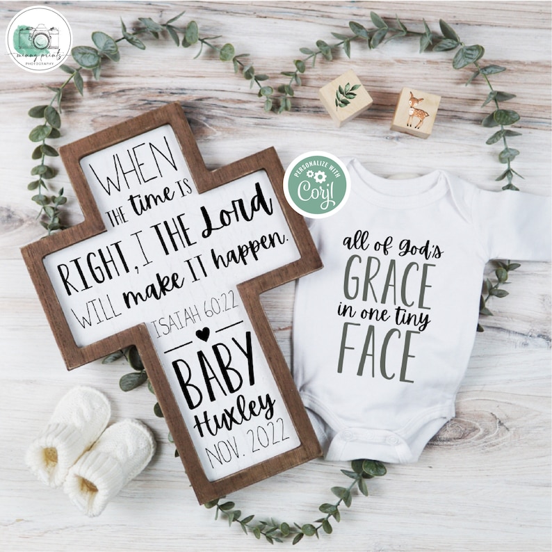 May include: A wooden cross with a white sign that reads "When the time is right, I the Lord will make it happen. Isaiah 60:22". Below the sign is a heart and the text "Baby Huxley Nov. 2022". A white onesie with the text "All of God's Grace in one tiny face" is next to the cross.