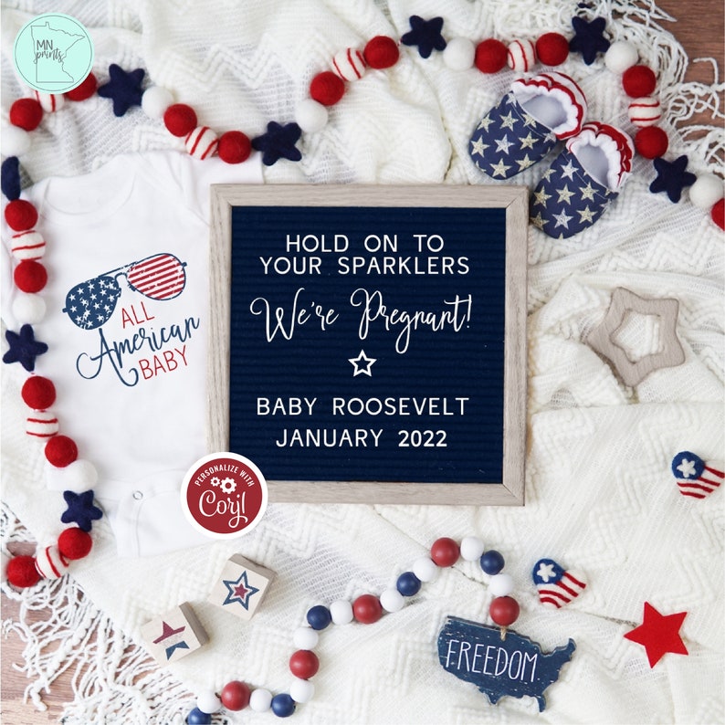 K&ouml;nnte beinhalten: Ein wei&szlig;er Strampler mit dem Text "All American Baby" und einer Sonnenbrillengrafik. Eine blaue und wei&szlig;e Buchstabenwand mit dem Text "Hold on to your sparklers We're Pregnant! Baby Roosevelt January 2022". Rot-wei&szlig;-blaue Babyschuhe, Holzkl&ouml;tze und eine Girlande aus rot-wei&szlig;-blauen Pompons.