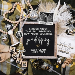 May include: A black letter board with the text "Enough about that ball dropping, how about something jaw dropping? Baby Clark July 202X". The board is surrounded by a variety of baby items, including a onesie, booties, a rattle, and a bottle.