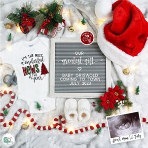 Op de afbeelding: Een kerstthema baby aankondiging met een witte romper met de tekst "It's the most wonderful news of the year" met een rood-zwarte geruite kerstboom. Een letterbord zegt "Ons grootste cadeau Baby Griswold komt naar de stad Juli 2023". Er is een kerstmanmuts, babyschoentjes, een kerstbal en een echobeeld.