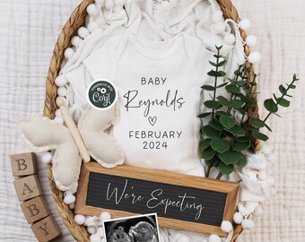 May include: A wicker basket filled with white pom poms, a white onesie with the text "Baby Reynolds, February 2024", a wooden sign that says "We're Expecting", a white butterfly toy, and a framed ultrasound image. The basket is on a white textured surface.