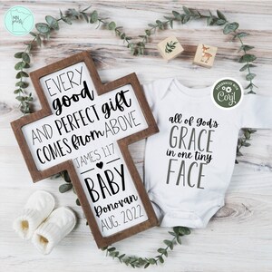 May include: A wooden cross with the text "Every good and perfect gift comes from above" and "James 1:17". Below the text is a heart and the words "Baby Donovan Aug. 2022". A white onesie with the text "All of God's Grace in one tiny face" is next to the cross.