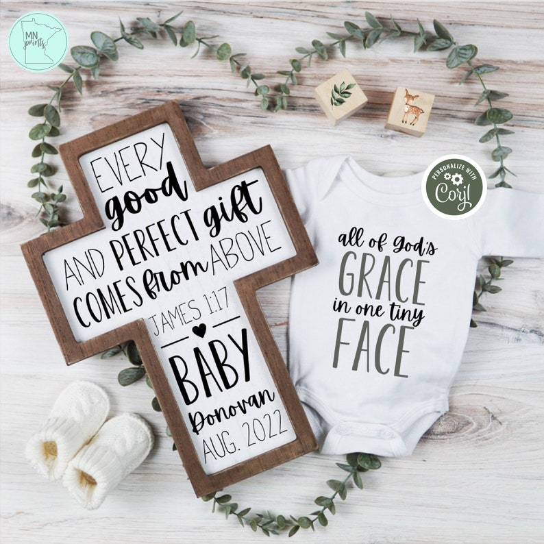May include: A wooden cross with the text "Every good and perfect gift comes from above" and "James 1:17". Below the text is a heart and the words "Baby Donovan Aug. 2022". A white onesie with the text "All of God's Grace in one tiny face" is next to the cross.